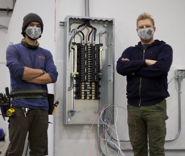 Two OHM electricians stand near a large industrial electrical panel.